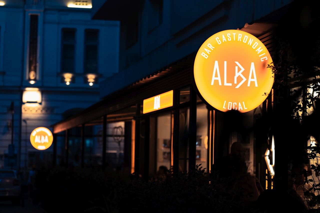 Cel mai nou restaurant cu specific românesc s-a deschis de curând pe Calea Victoriei, lângă Biserica Albă, în polul de turism al orașului, dar și pe o stradă cu tradiție a capitalei.  Despre restaurantul Alba Bar Gastronomic Local Restaurantul a preluat numele de la biserica emblematică lângă care se află, dar și pentru că albul transmite ideea de curat și simplu, exact spiritul pe care îl exprimă și sloganul lor: „atât de simplu, atât de bun”. În spațiul respectiv au funcționat timp de 10 ani sub un alt nume și concept, dar la sfârșitul lunii iulie, un incendiu nefericit i-a forțat să o ia de la capăt. Datorită eforturilor uriașe ale întregii echipe, au reușit să reconstruiască totul în doar patru săptămâni. Sub noua denumire și noul concept, restaurantul funcționează din 1 septembrie 2025. Restaurantul are 50 locuri în interior și 50 locuri pe terasă, atunci când vremea o permite. Designul spațiului, atât estetic cât și funcțional, inclusiv mobilierul, a fost creat și realizat de echipa Arhart Creative Studio. “Conceptul „Nostalgia modernismului integrată în prezent – atât de simplu, azi” se reflectă în ansamblul arhitectural de pe peretele de la răsărit, conceput ca un altar, care sugerează punctul de pornire și importanța rădăcinilor. Partea centrală reprezintă orașul într-un zbucium de forme, cu o cromatică vibrantă, încadrată de menajeria de coloane care captează lumina necontrolat, generând un dramatism sobru ce creează conexiunea între pământ și cer”, detaliază  Bogdan Mesteru, creative director. Pentru că „unora le place jazz-ul”, vineri și sâmbătă de la 19:30 și duminica la brunch, au artiști live cu voce, pian, vioară și saxofon. În restul zilelor, oaspeții se pot bucura de muzică românească, de la șlagăre clasice, la piese moderne, degustări de vin alături de cramele partenere și de expoziția permanentă de lucrări ale artistului român Mircia Dumitrescu. Despre meniul Alba Bar Gastronomic Local Restaurantul are un specific local, românesc, construit în jurul ideii de nostalgie. Își doresc să readucă în farfurie gusturile cu care mulți dintre noi am crescut, prezentate într-o formă modernă. Meniul a fost conceput de întreaga echipă. A fost un proces colectiv, în care fiecare a venit cu propriile nostalgii și amintiri. După o cercetare atentă a pieței, au ales să lucreze cu ingrediente de la mici producători locali, de încredere. Chef Nicușor Stoican și Chef Mihai Androne pun în valoare întreaga echipă din bucătărie, coordonând-o. Vedetele meniului sunt ciorba de burtă, jumările calde, micii mici, ficatul de vițel și coliva cu blat de biscuiți, însă puteți găsi aici și prăjituri pe care ni le făceau mama sau vecina, precum albă-ca-zăpada ori salamul de biscuiți.  Alba Bar Gastronomic Local